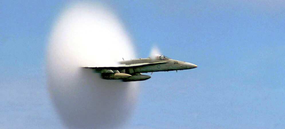 Sky surrounded by hazy clouds with a military airplane flying three quarters out of a large, vertical oval cloud.