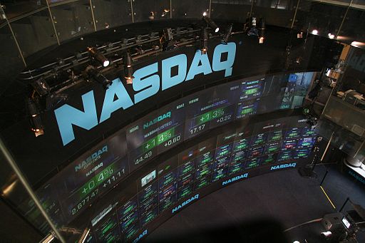 Birdeye view of the NASDAQ trading floor, with spotlight lighting fixtures atop a banner with NASDAQ. Below the banner are two large stacked curved monitors with stock information in numbers and stock ticker symbols.