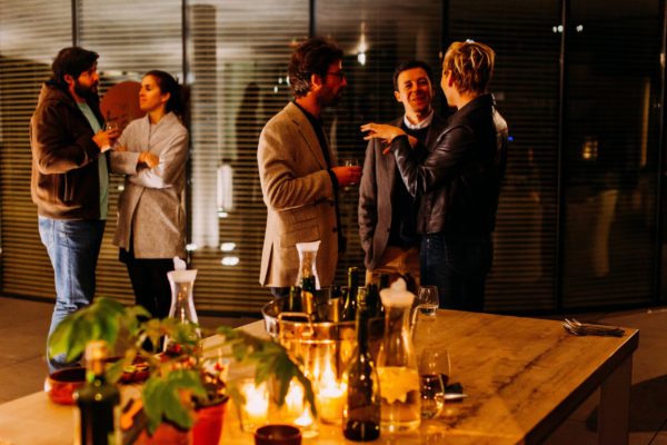 Social gathering showing a man and woman talking to each other in the back left, and a group of three people conversing together in the center. A table is in the foreground, with carafes of water and wine bottles.