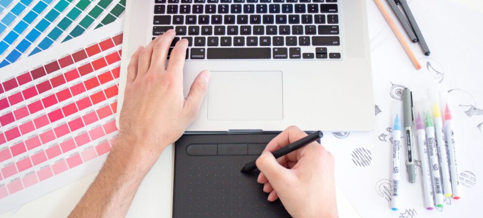 Desk with laptop's keyboard. One hand is resting on the keyboard, the other is drawing on an electronic pad. Color pens rest on the right of the keyboard, several colorful paint swabs are on the left.