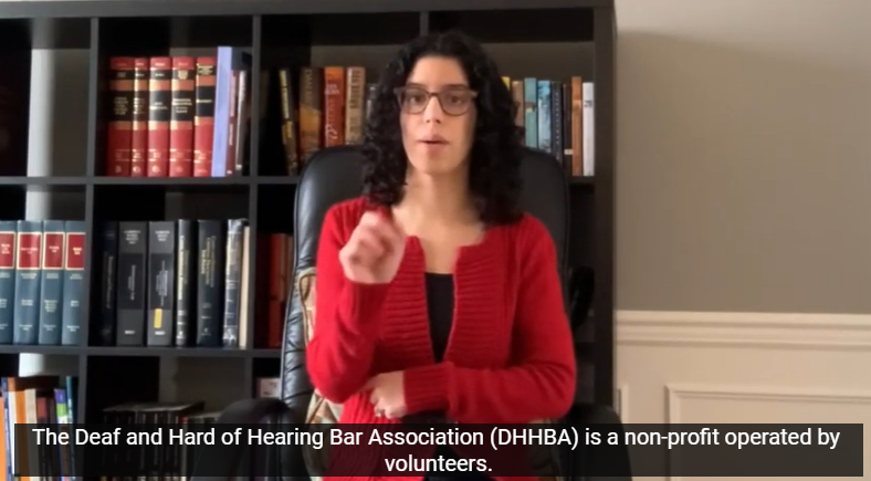 Zainab Alkebsi, DHHBA President, is wearing a red blazer over a black top. She has black curly hair with brown glasses. She is sitting in her home office in front of a black bookcase and two-toned grey/white walls.